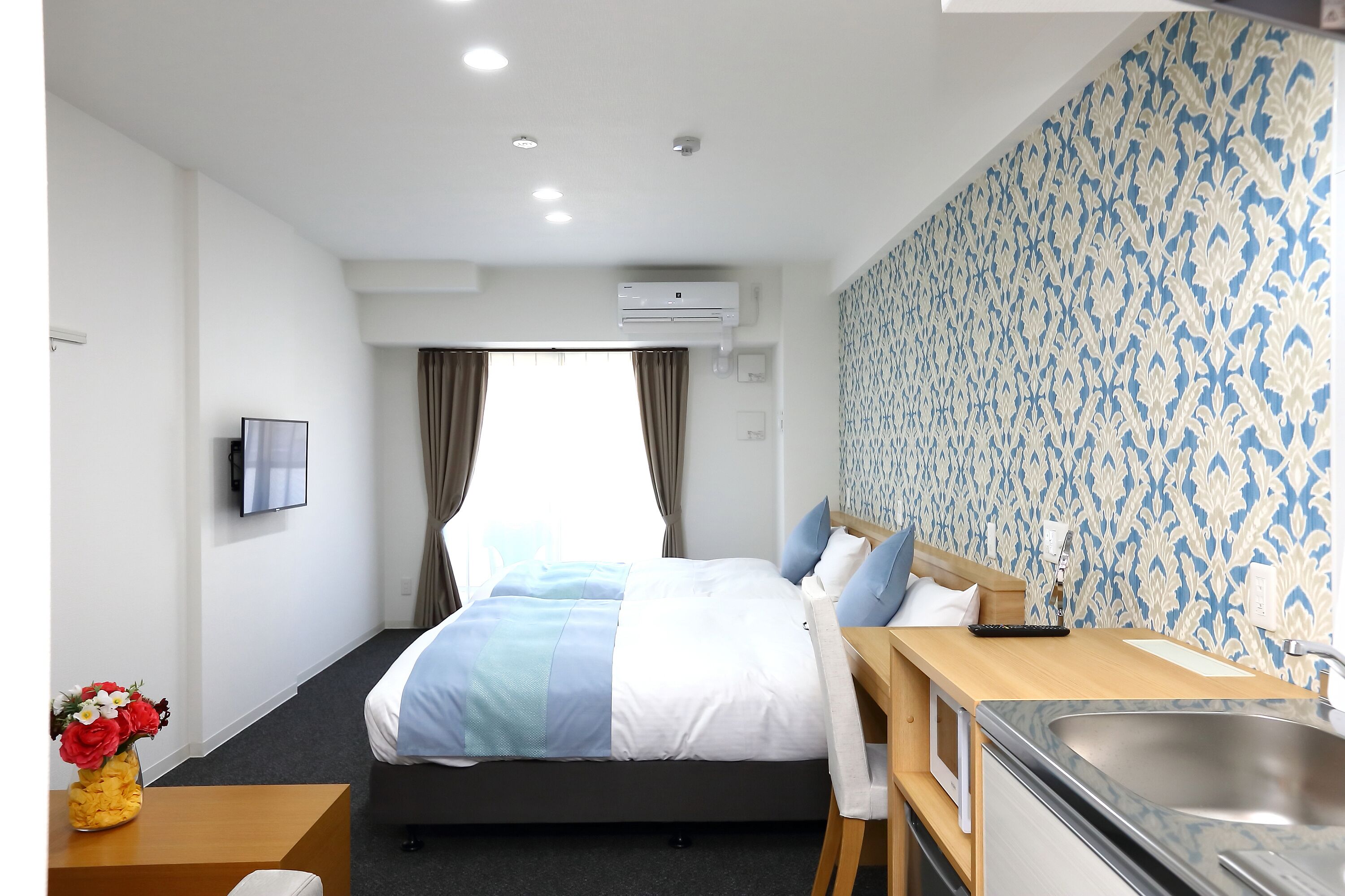 A hotel room with a bed, a desk with a sink, a flat-screen TV, and an air conditioning unit.