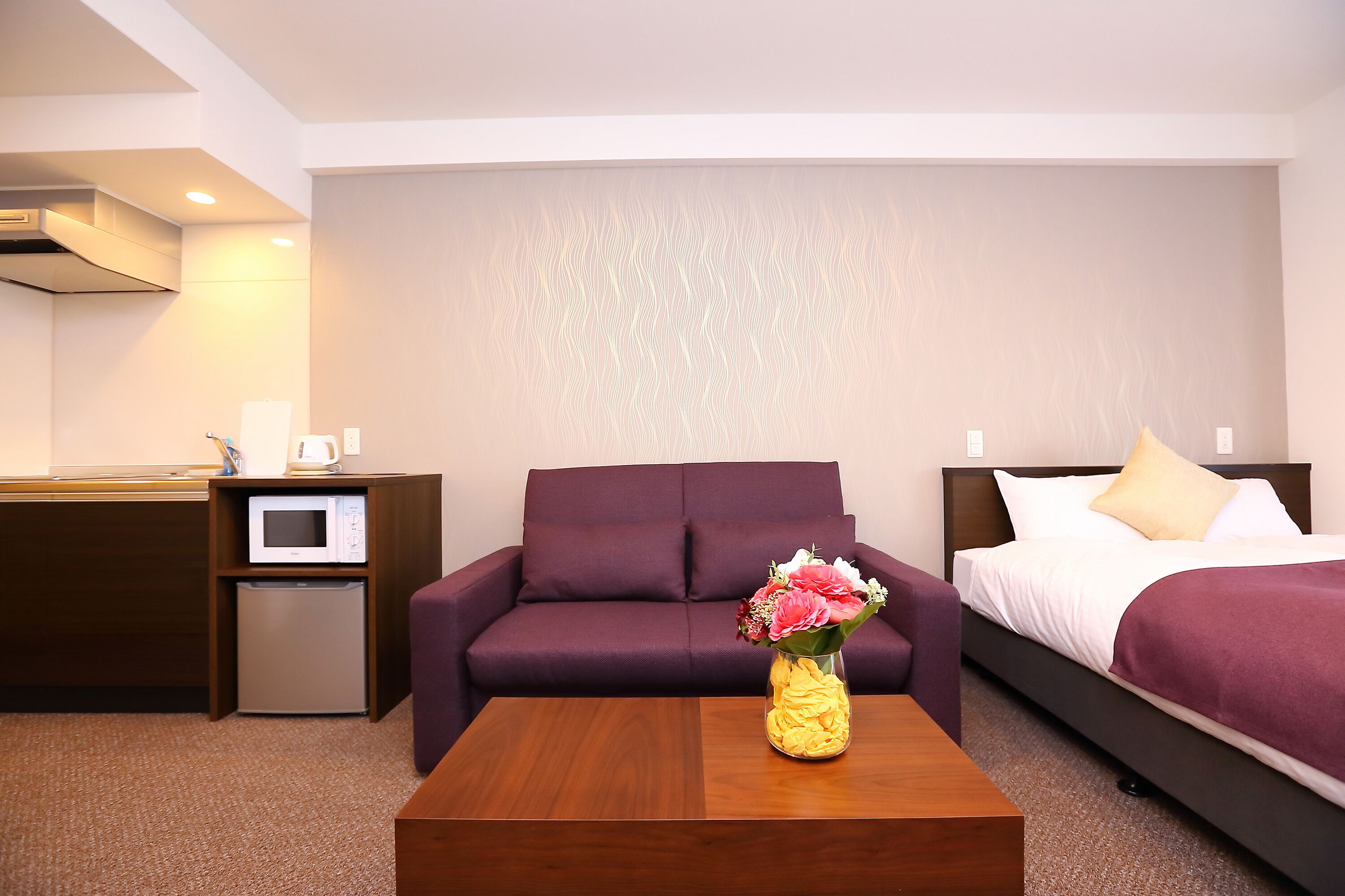 A hotel room with a purple sofa, a bed with a purple bedspread, a wooden coffee table with a vase of flowers, a microwave, and a small refrigerator.