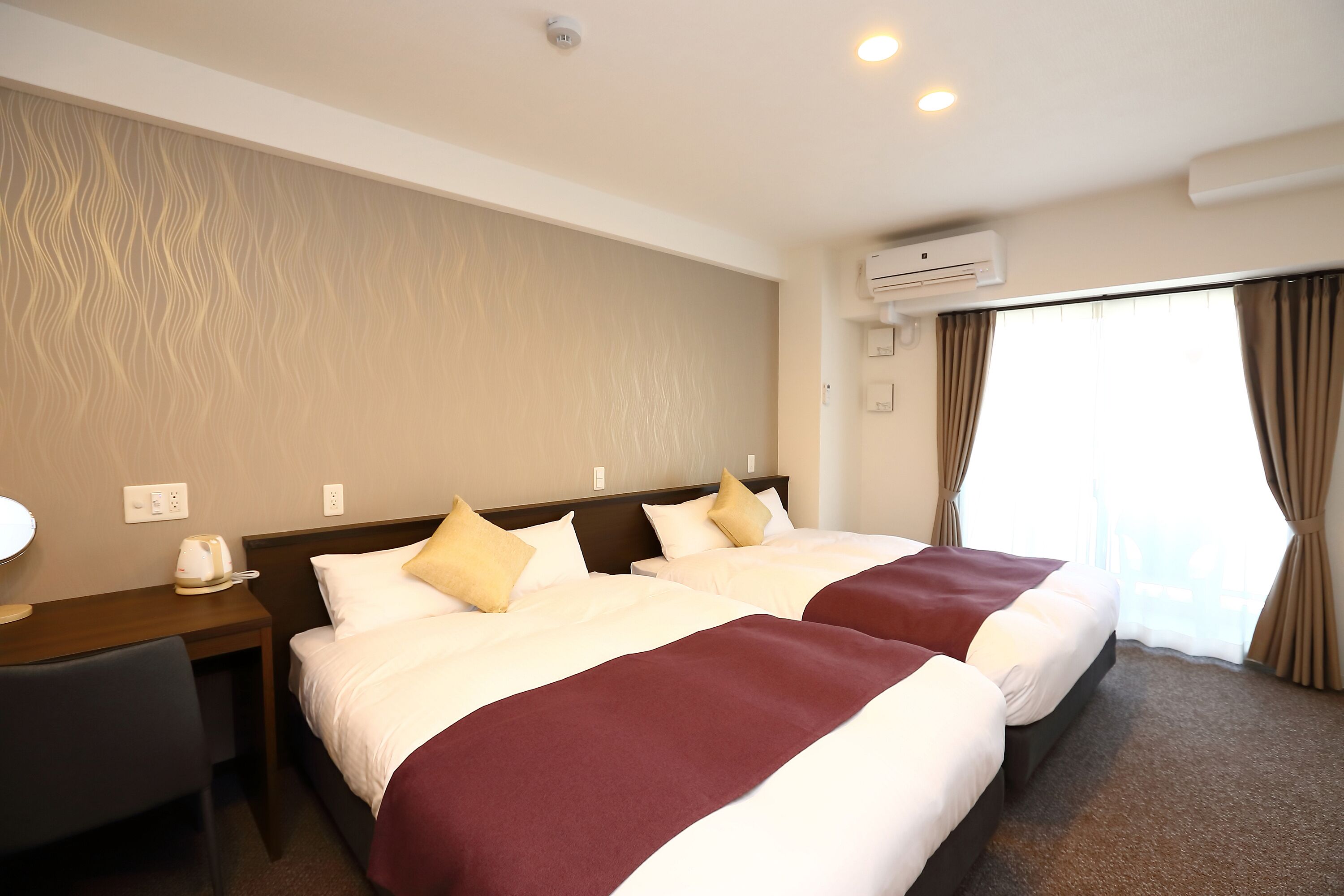 A hotel room with two beds, a desk, a chair, and an air conditioning unit.