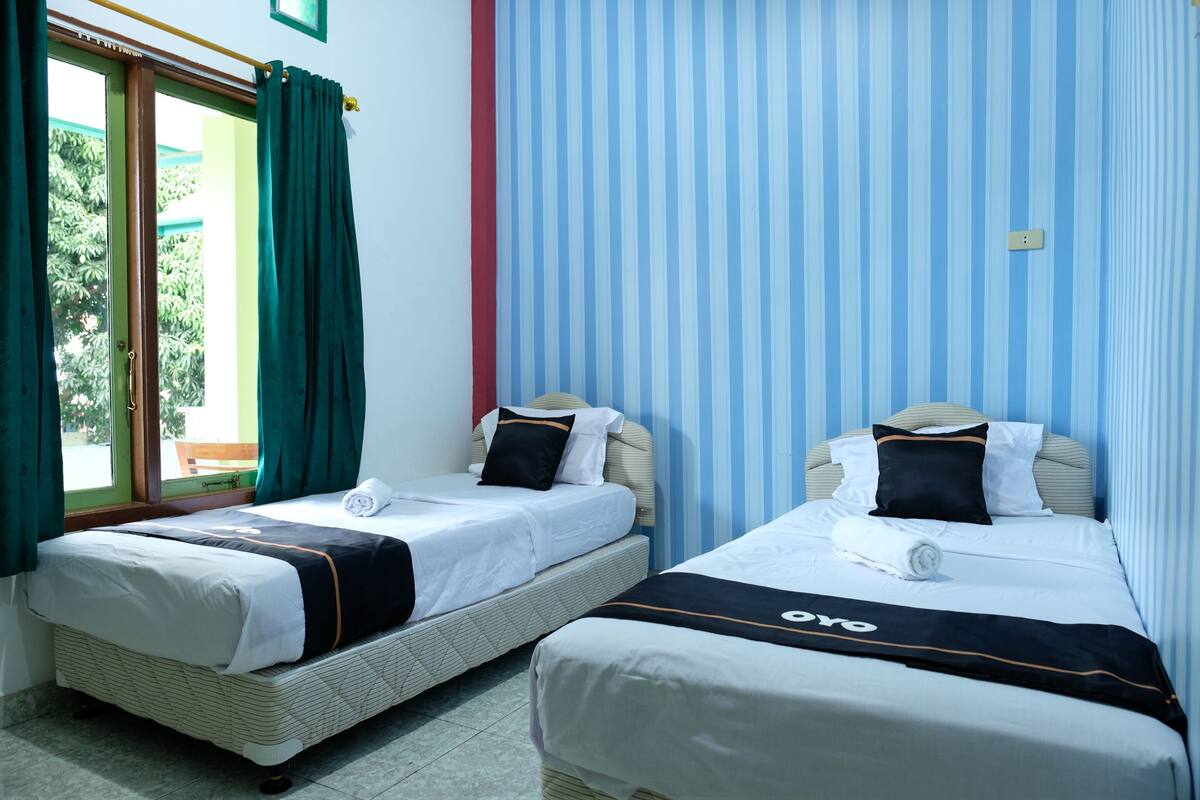 Twee enkele bedden met witte beddengoed en zwarte accenten, een raam met groene gordijnen en een blauwe muur met strepen.