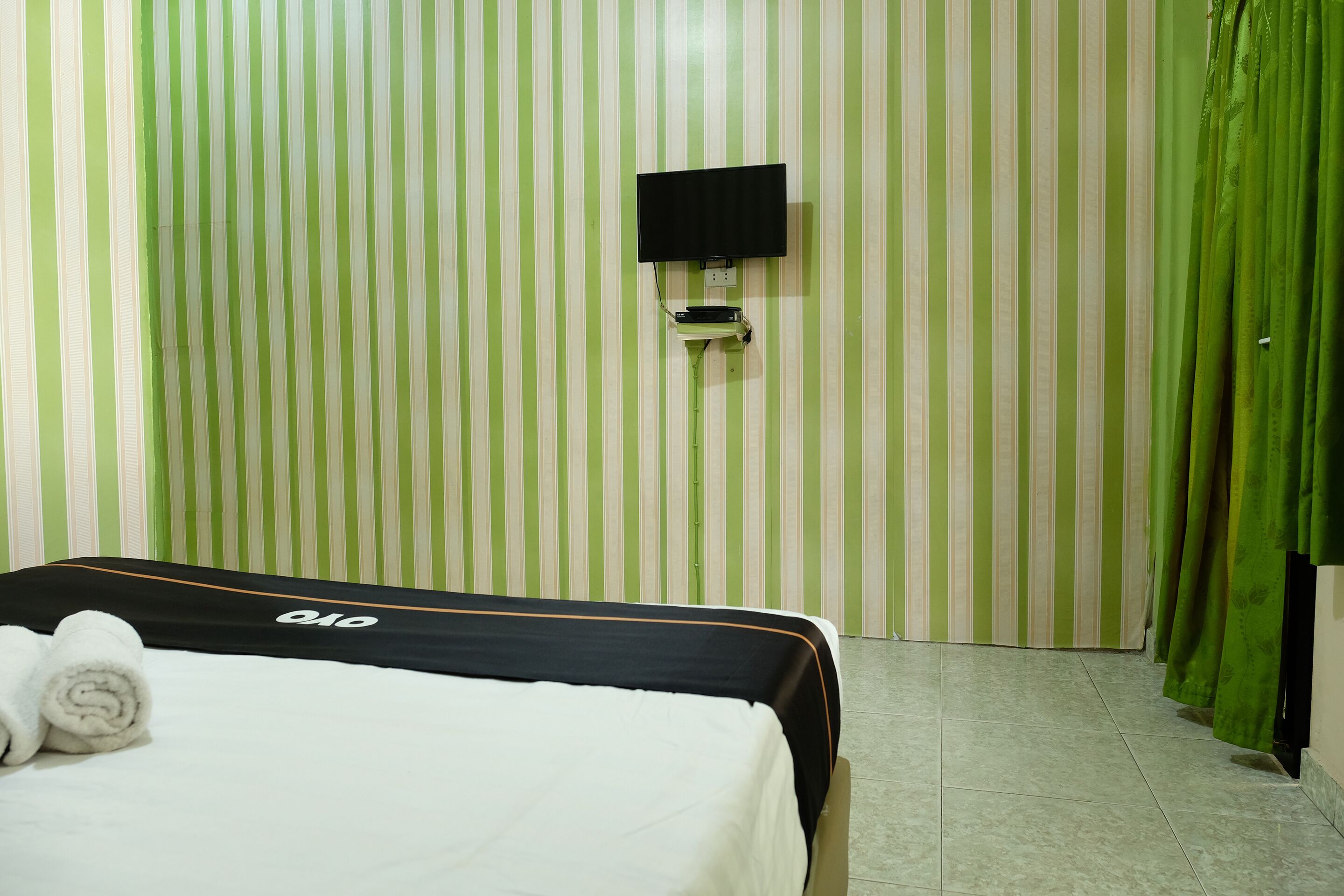 Una habitación de hotel con pared verde y blanca a rayas, un televisor de pantalla plana montado en la pared, una cama con colcha blanco y negro y cortinas verdes.