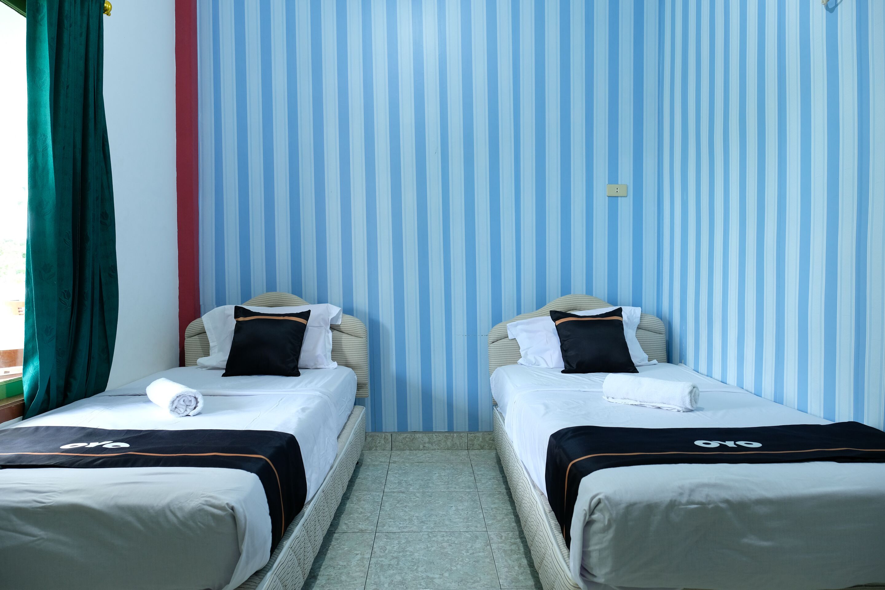 Une chambre d’hôtel avec deux lits simples, un papier peint à rayures et un rideau vert.