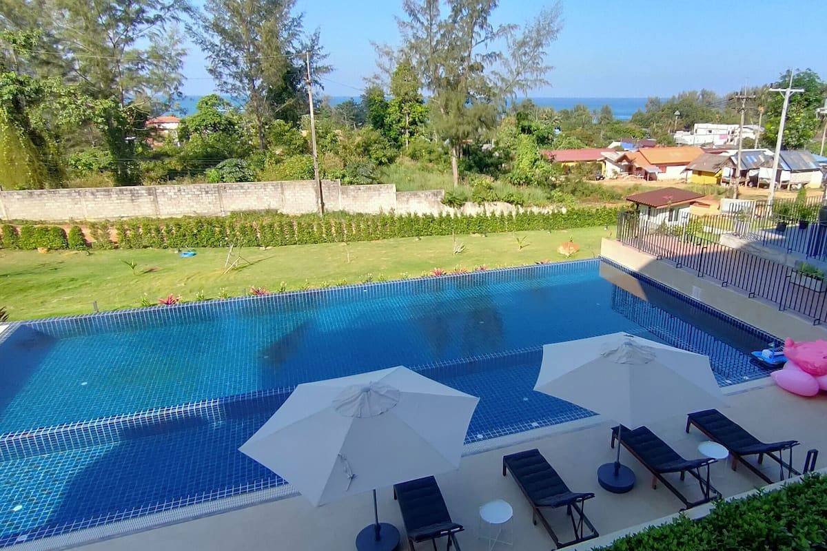 A rectangular swimming pool with a tiled deck, surrounded by lounge chairs and umbrellas, with a view of a residential area and trees in the background.
