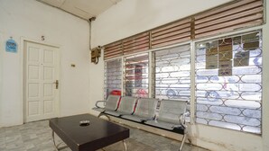 Een wachtruimte met een tafel, stoelen, een deur met het nummer 5 en een raam met metalen tralies.