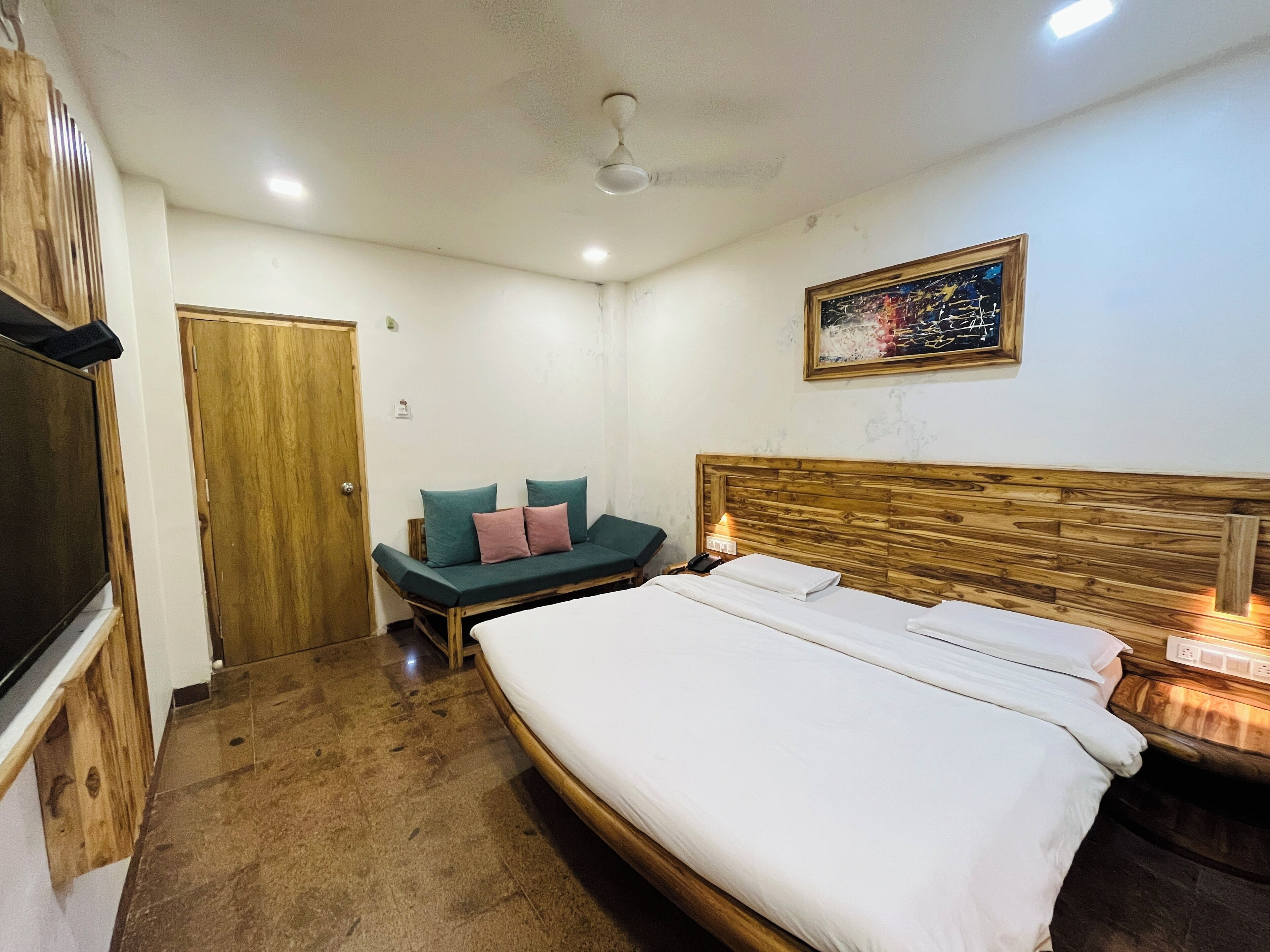 Ein Hotelzimmer mit einem großen Bett, einem Holzkopfende, einer kleinen Couch mit Kissen, einer Holztür und einem Bild an der Wand.