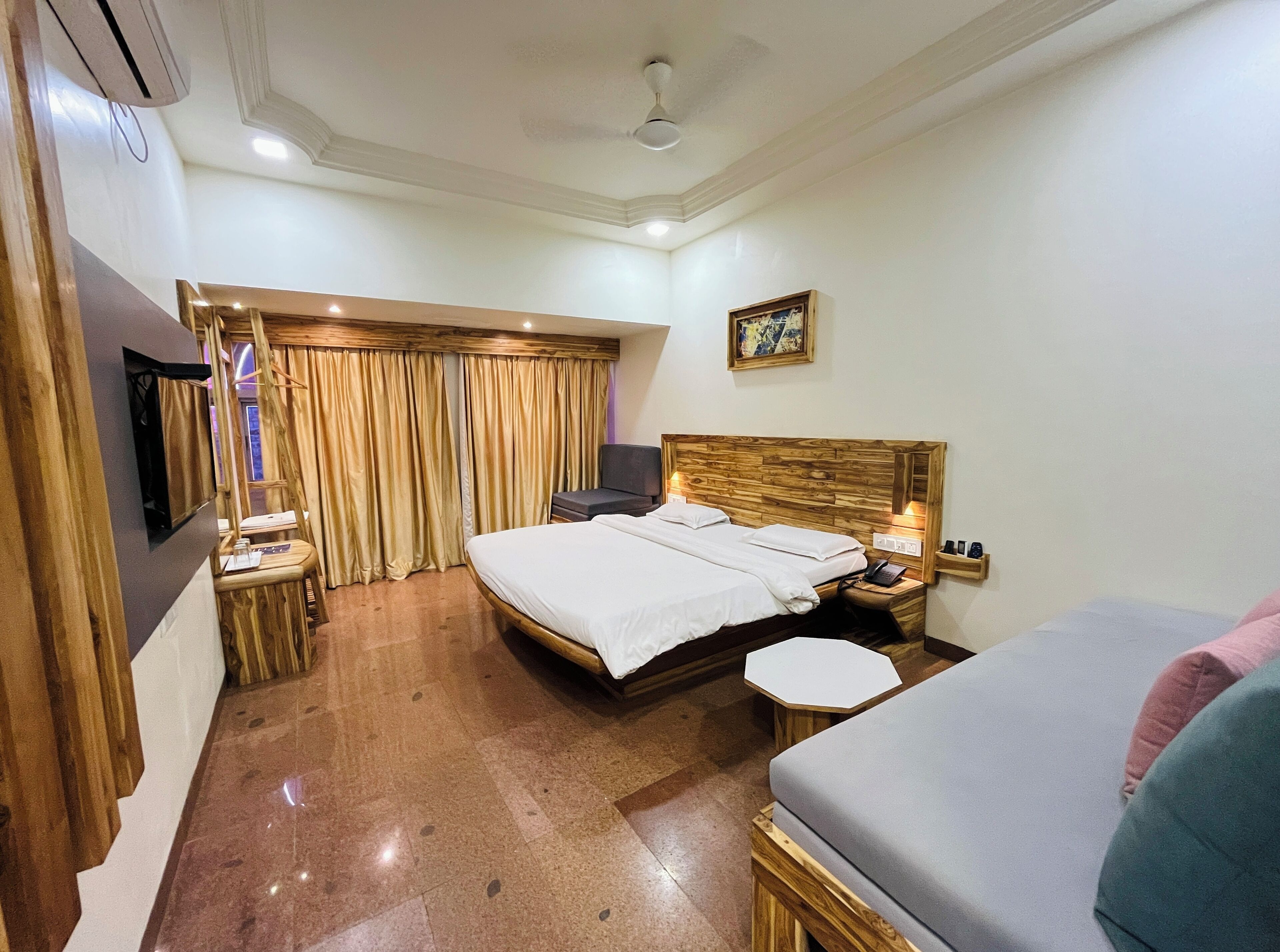 Habitación de hotel con cama de cabecera de madera, una mesita de noche pequeña, televisor montado en la pared y una ventana con cortinas.