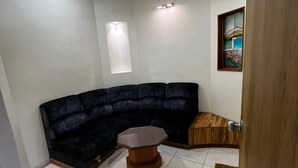 A room with a dark blue sectional sofa, a wooden coffee table, a ceiling fan, recessed lighting, a window with a view, and a framed artwork on the wall. - Hotel Shripad Continental (Amravati)