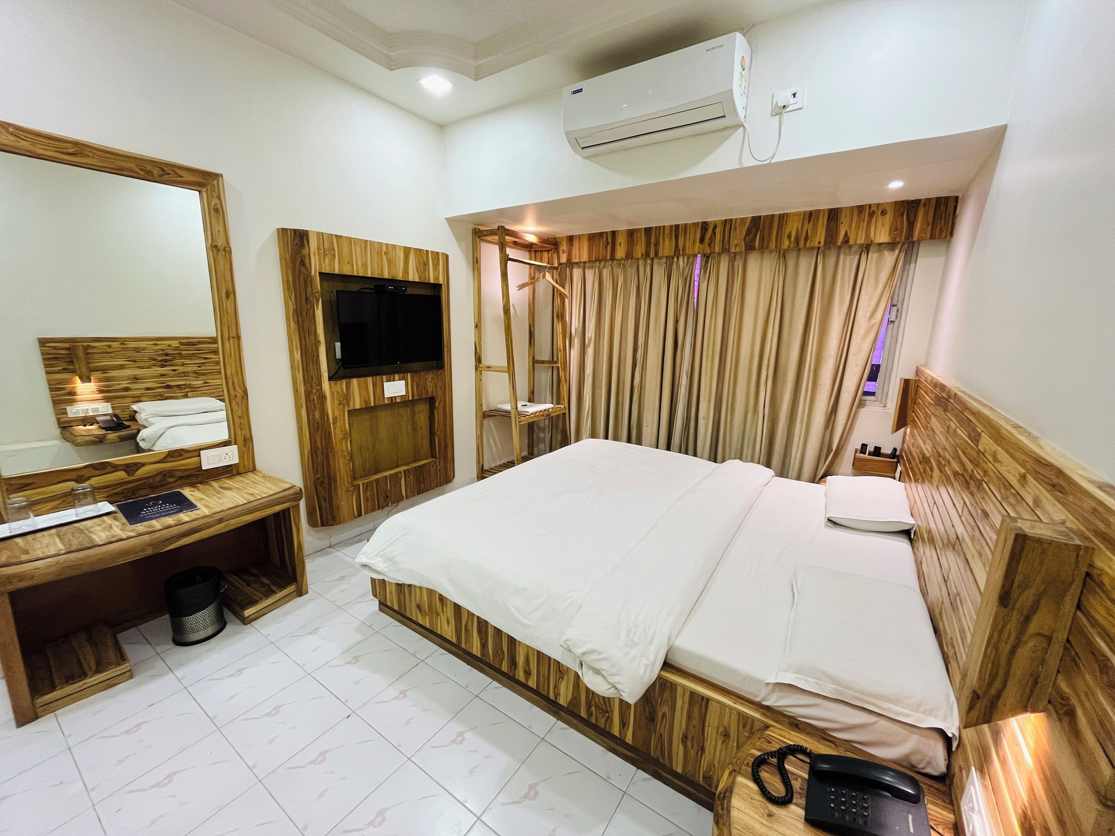 A hotel room with a wooden headboard, a large mirror, a flat-screen TV, a bedside table with a phone, and a window with curtains.