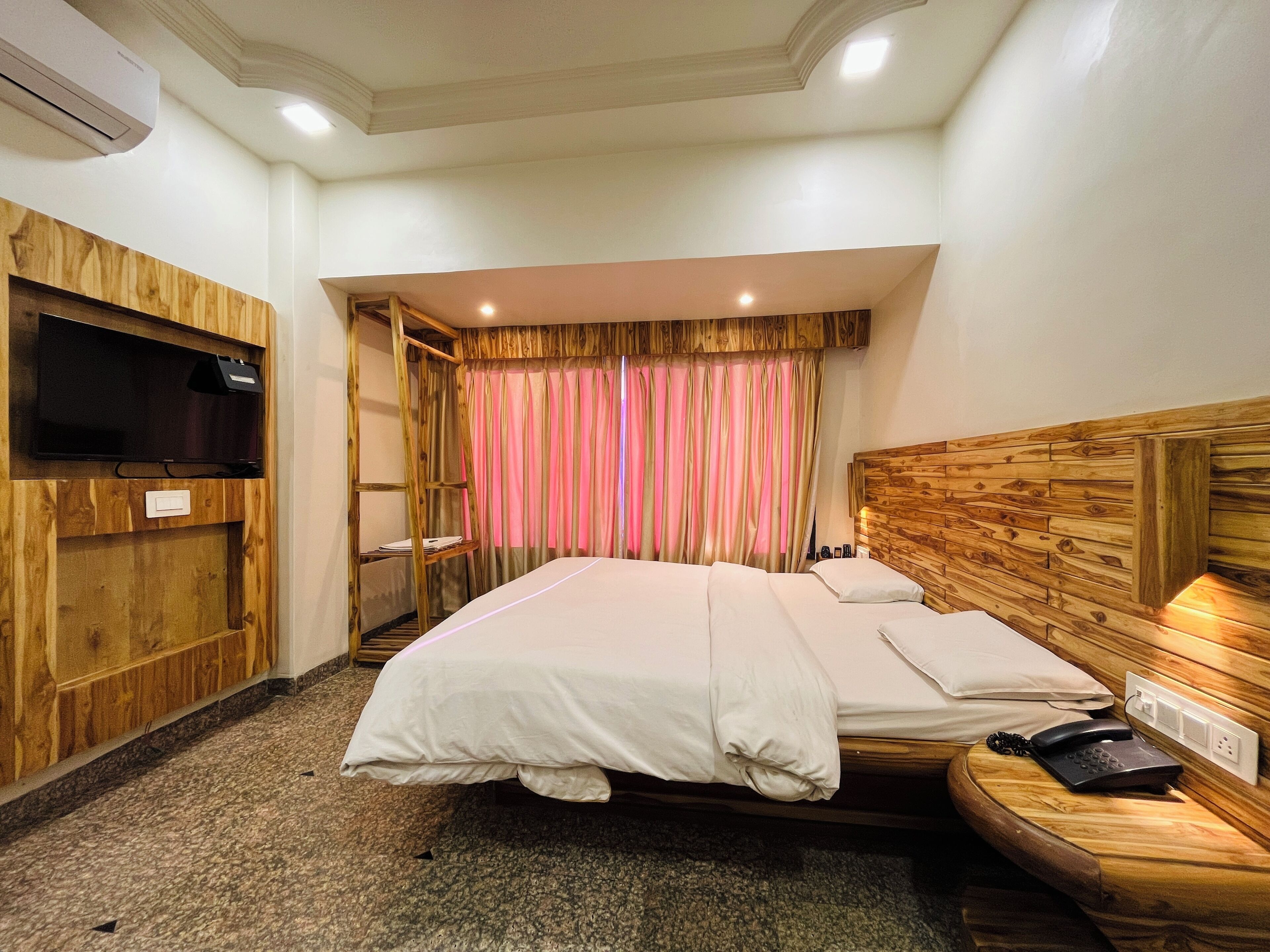 Una camera d'albergo con una testiera in legno, un letto con lenzuola bianche, una televisione a parete e un telefono sul comodino.