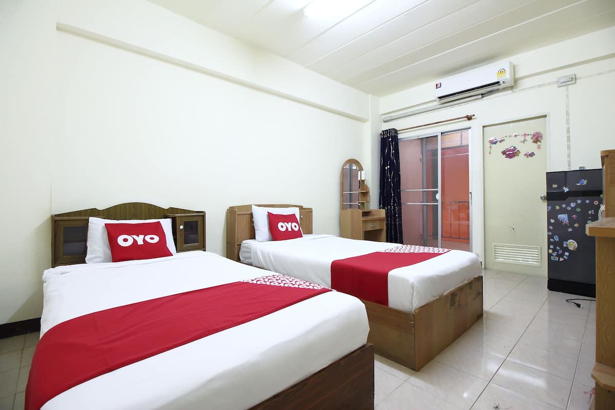 Habitación de hotel con dos camas, cada una con cabecero de madera, ropa de cama blanca y detalles en rojo. Se observa un armario de madera, una mesita y un equipo de aire acondicionado en la pared.