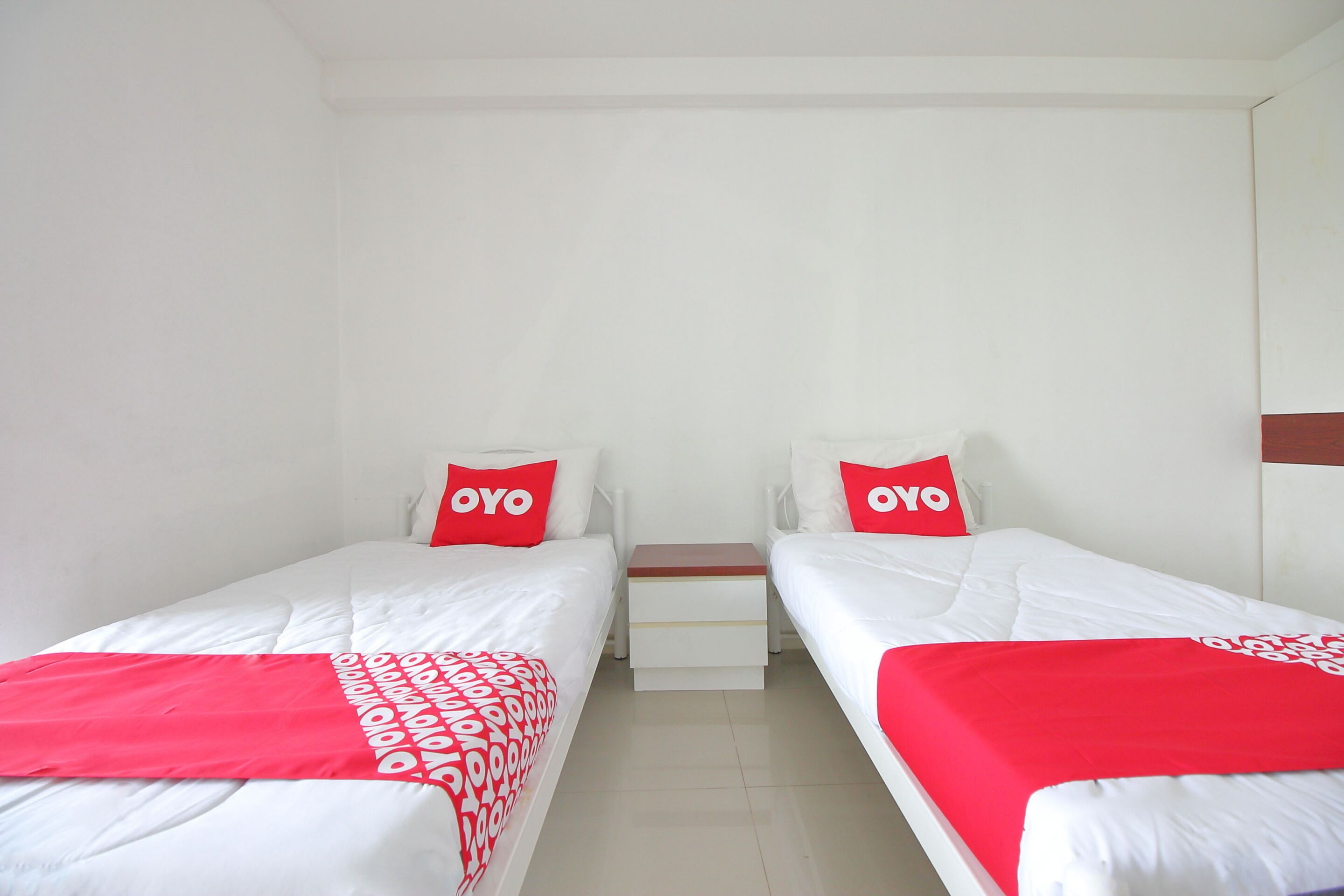 A hotel room with two single beds, each with a red pillow and a white bedspread.