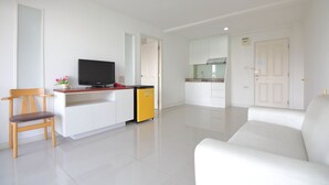 Ein modernes Wohnzimmer mit einem weißen Sofa, einem Holzstuhl, einem Fernseher und einer kleinen Küchenzeile mit Schränken und einer gelben Mülltonne.