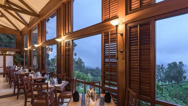 A restaurant with wooden furniture and large windows offering a view of the outdoors. - Club Mahindra Mount Serene Resort in Munnar (Devikolam)