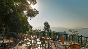 An outdoor dining area with tables and chairs, surrounded by greenery and a view of distant mountains. - Club Mahindra Mount Serene Resort in Munnar (Devikolam)