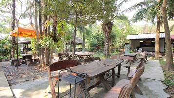 An outdoor dining area with wooden tables and benches, surrounded by trees and covered by a canopy.