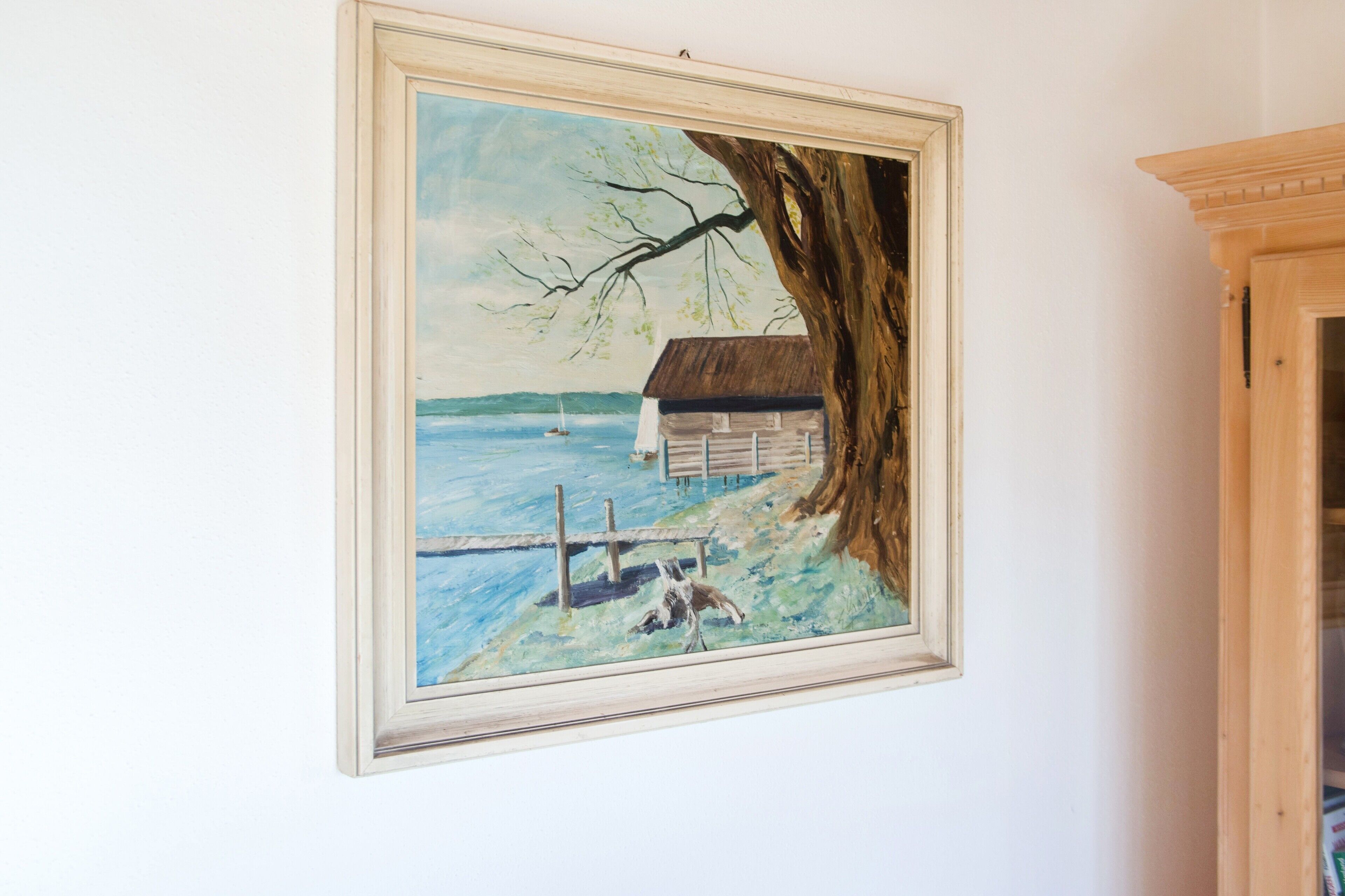 A framed painting of a coastal scene with a house, a tree, and a dock.
