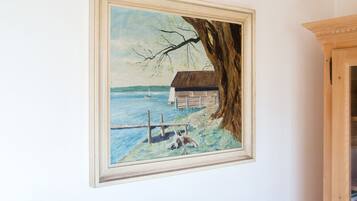 A framed painting of a coastal scene with a house, a tree, and a dock.