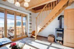 Un escalier en bois menant à une mezzanine, une grande porte-fenêtre coulissante donnant sur une terrasse et un poêle à bois.