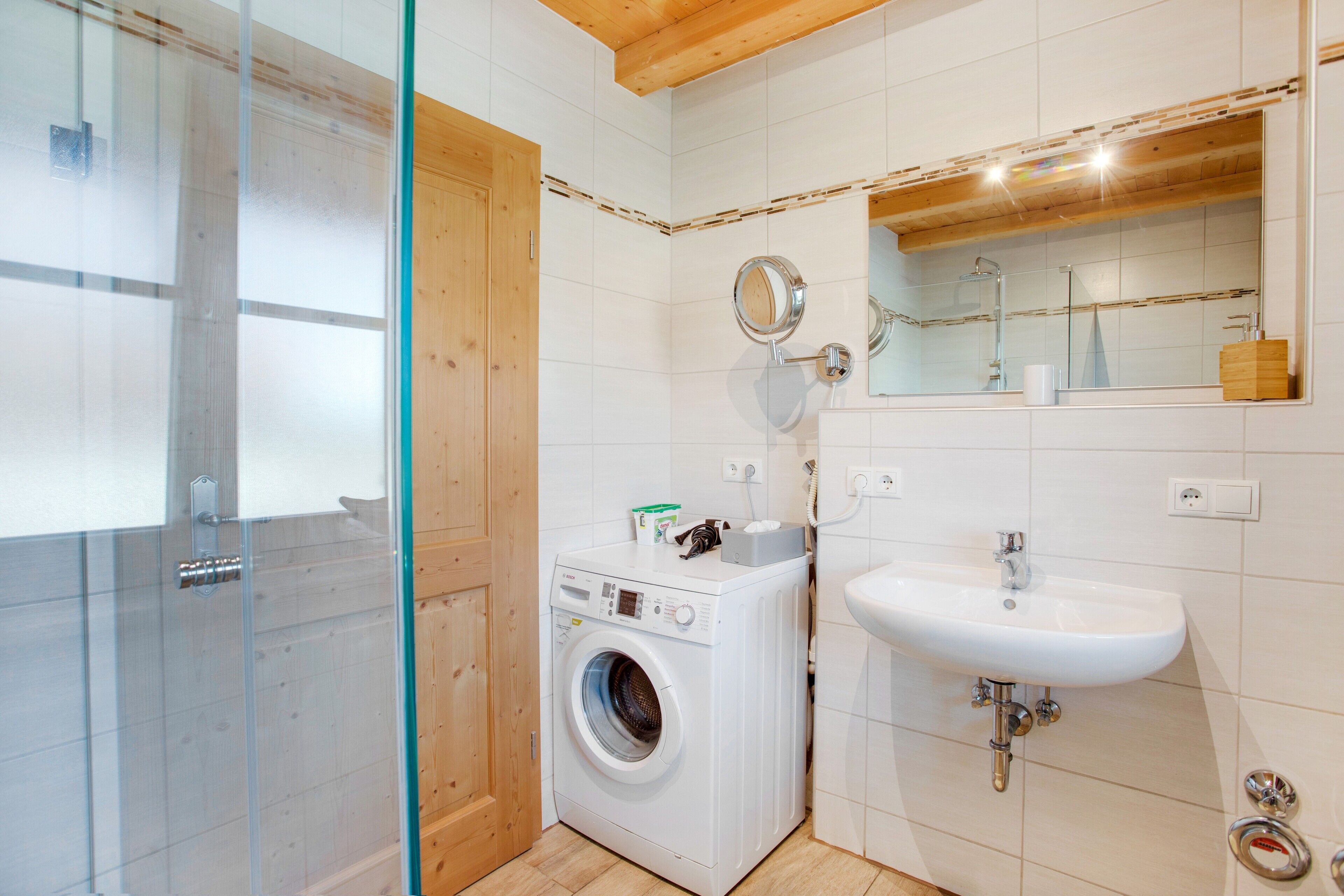 Un baño con mampara de vidrio, lavadora, lavamanos y espejo.