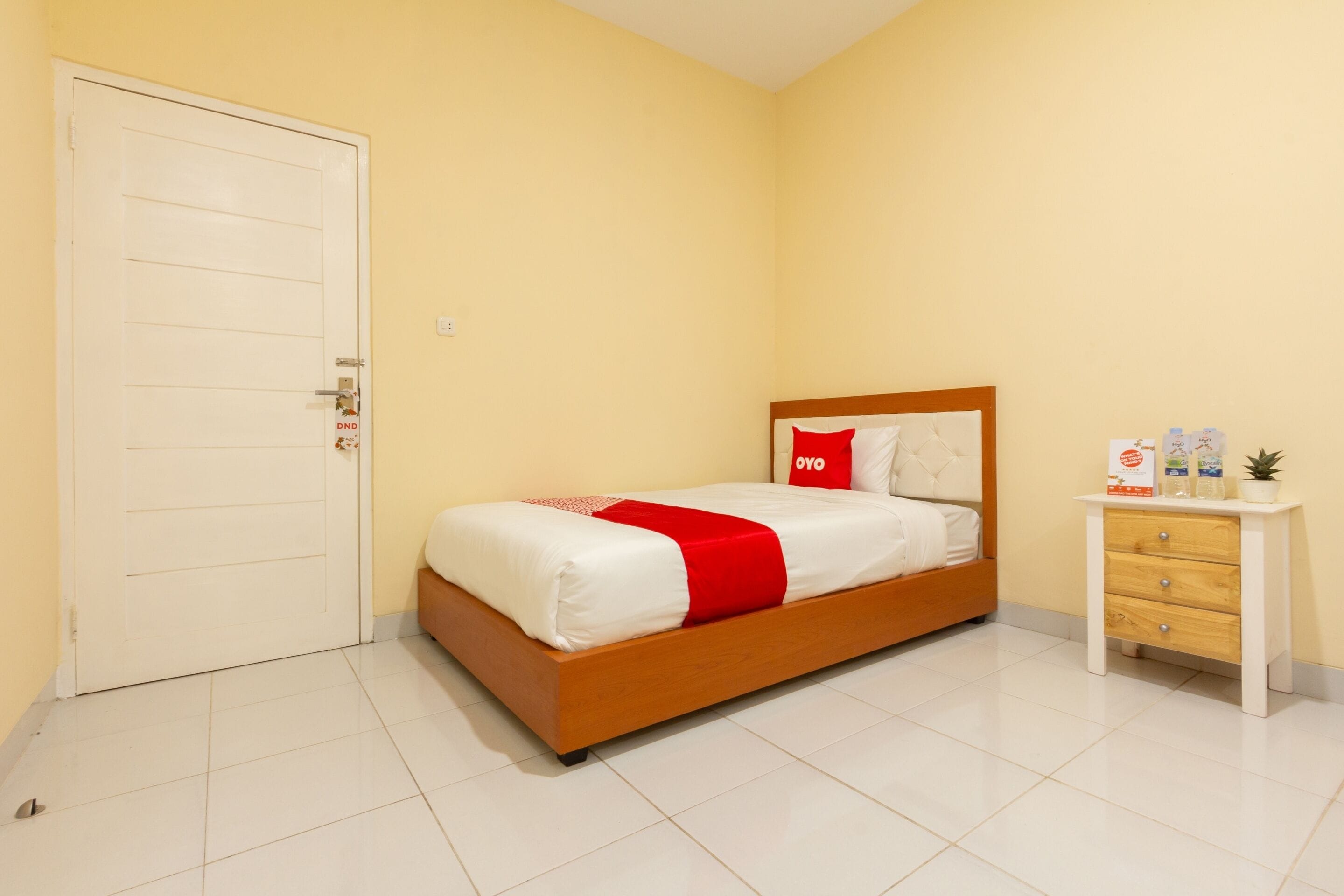 Un dormitorio con cama de madera, ropa de cama blanca, almohada roja, mesita de noche con planta y puerta con pomo.