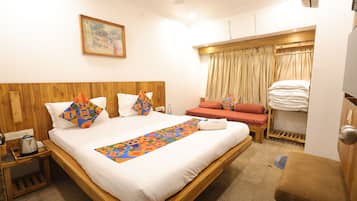 A hotel room with a large bed, a wooden headboard, a painting on the wall, a window with curtains, and a small seating area with a bench.