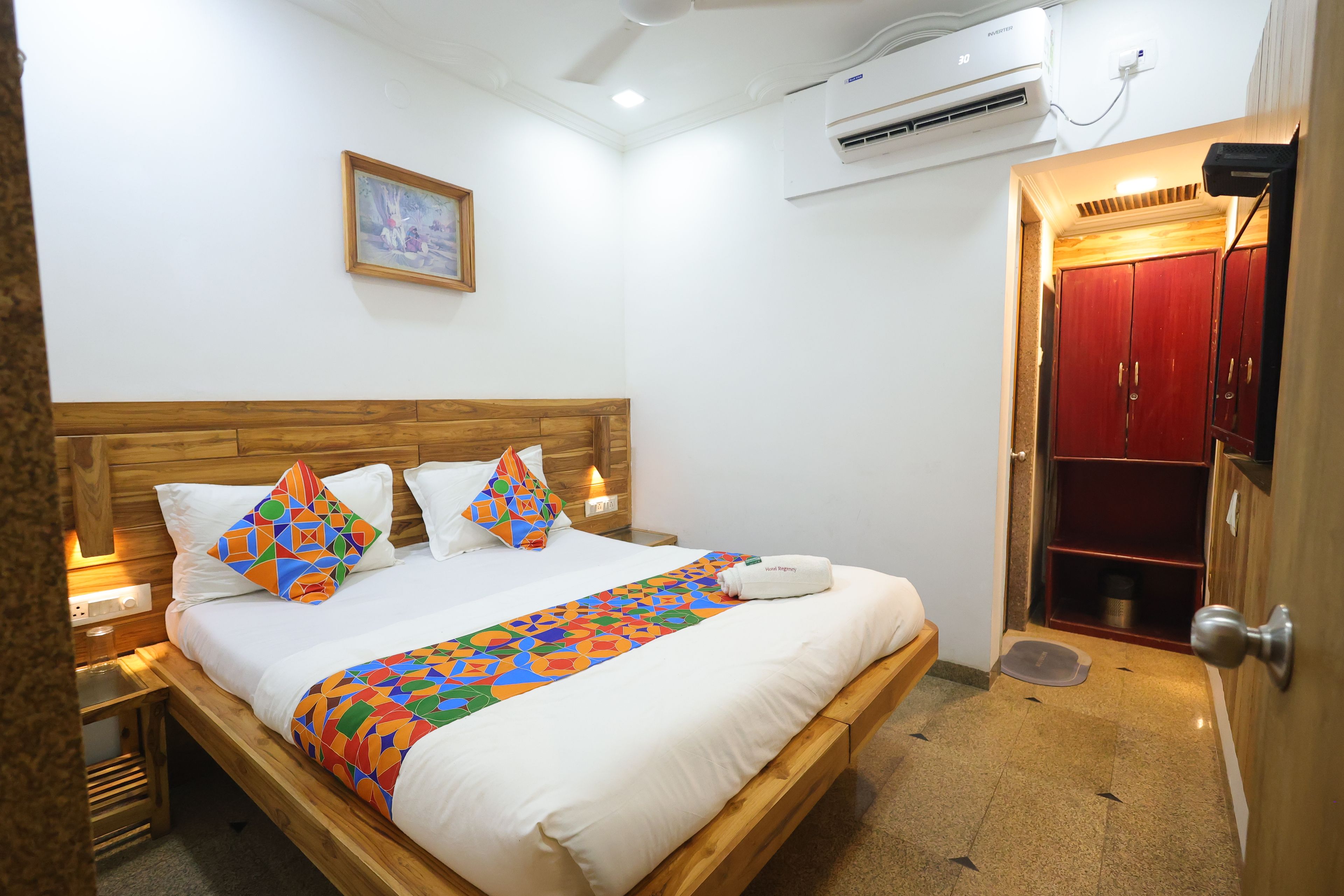 A hotel room with a wooden bed, white bedding, a colorful throw, a painting on the wall, an air conditioner, and a red wardrobe.