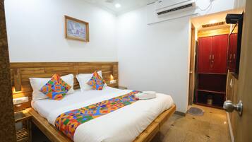 A hotel room with a wooden bed, white bedding, a colorful throw, a painting on the wall, an air conditioner, and a red wardrobe.