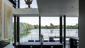 A modern dining area with a large table and chairs, a vase with flowers, and a view of a lake through large windows. - Gorgeous Lakeside Villa with wellness SPA and 8 seater cinema (Lechlade-on-Thames)