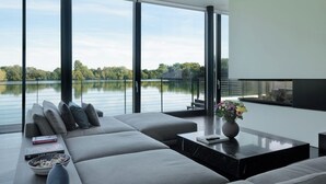 A modern living room with a large window overlooking a lake, grey sofas, a coffee table, and a fireplace. - Gorgeous Lakeside Villa with wellness SPA and 8 seater cinema (Lechlade-on-Thames)