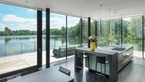 A modern kitchen with a large island, bar stools, and a view of a lake. - Gorgeous Lakeside Villa with wellness SPA and 8 seater cinema (Lechlade-on-Thames)