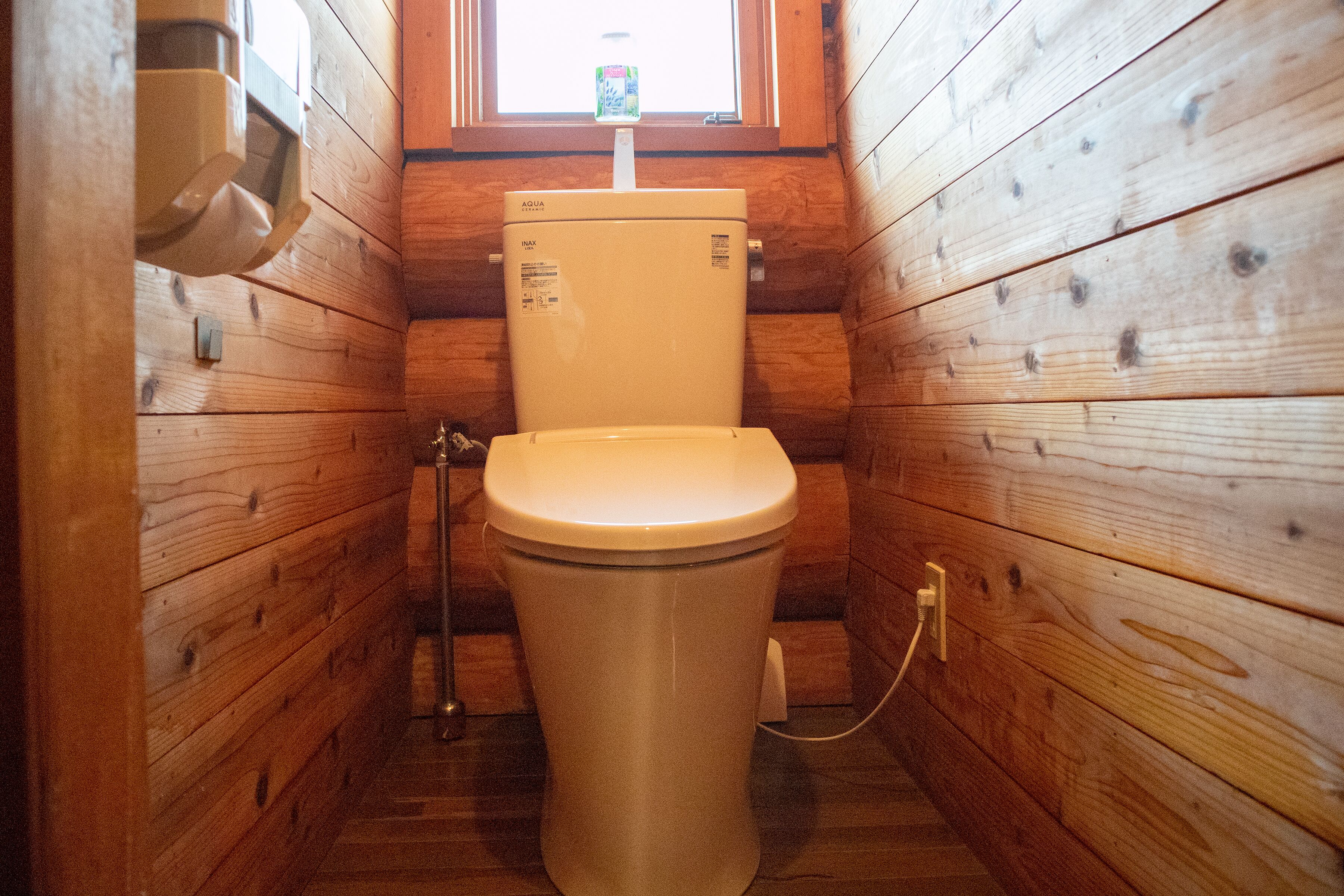 A small, wooden-paneled bathroom with a toilet, a window above the toilet, and a visible water heater.