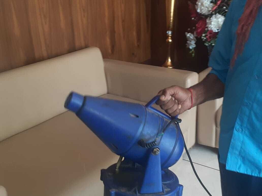 Une personne utilise un sèche-cheveux bleu sur un canapé beige, dans une pièce aux murs en bois et où se trouve un vase décoratif rempli de fleurs.