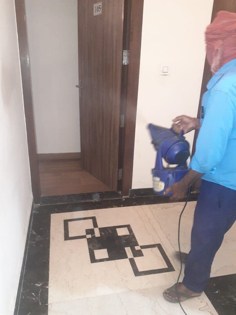 Una persona de pie en un pasillo con piso estampado, una puerta con un número y un objeto azul en el suelo.