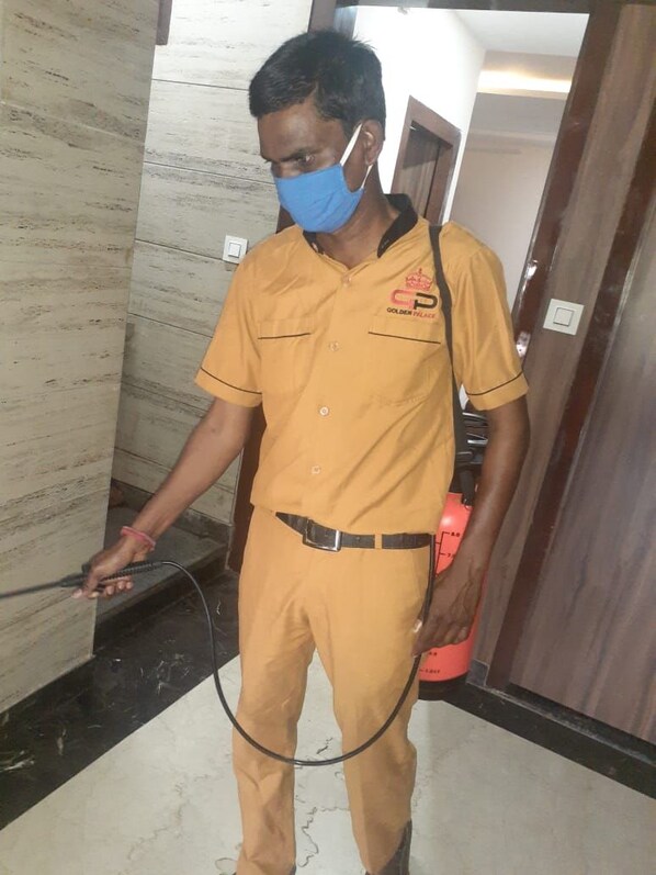 Una persona con uniforme amarillo y un logotipo, sosteniendo una manguera y un recipiente rojo, de pie en un pasillo con piso de mármol y puerta de madera.