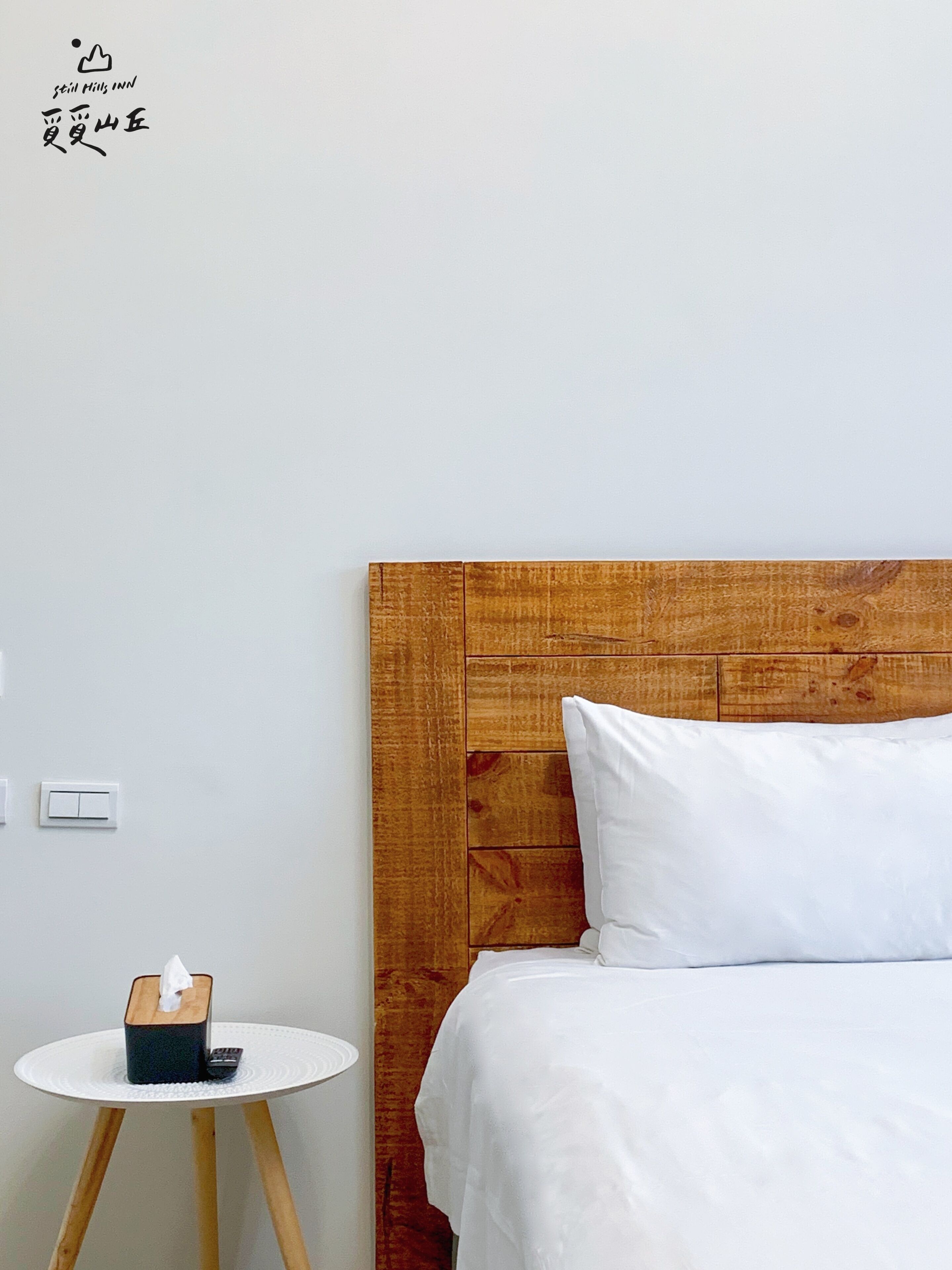 Habitación de hotel con cabecera de madera, ropa de cama blanca, una mesita de noche con caja de pañuelos y una pequeña mesa redonda.