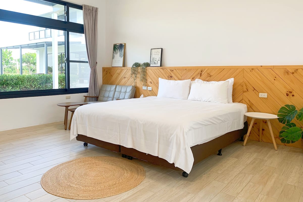 Un dormitorio moderno con una cama grande, cabecera de madera, una mesita auxiliar pequeña, un tapete redondo y vistas a zonas verdes.