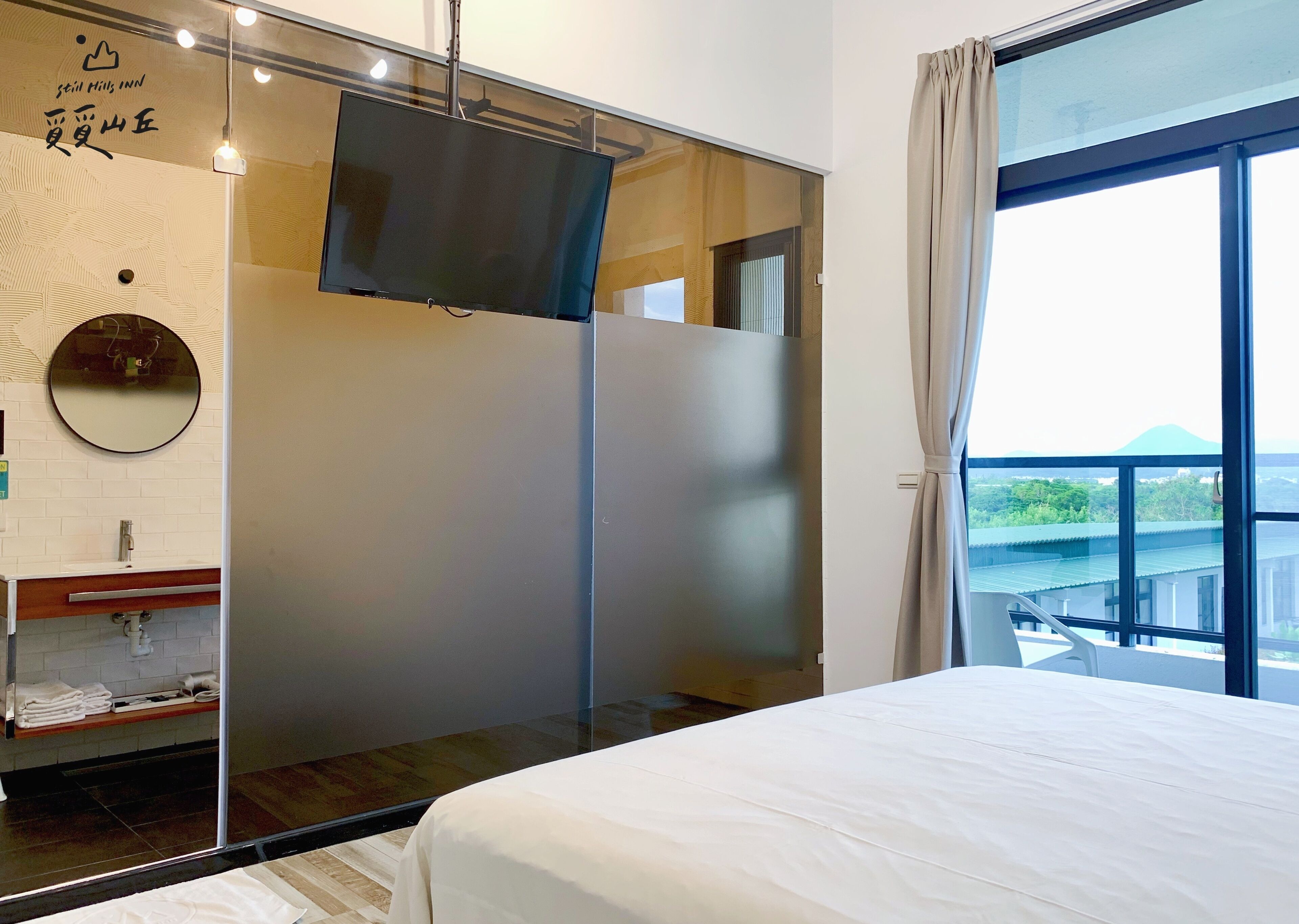 A modern hotel room with a large bed, a TV mounted on a glass wall, a bathroom with a mirror and sink, and a balcony with a view of the mountains.
