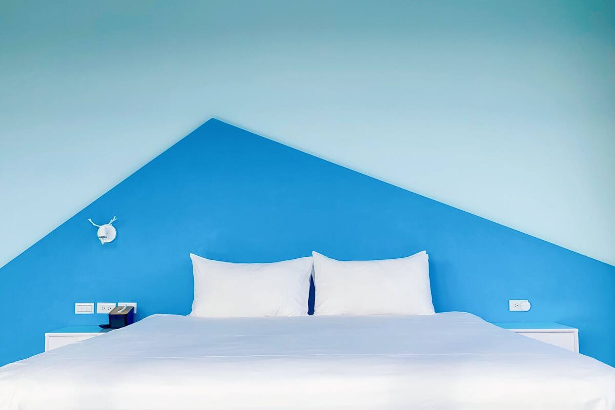 Un dormitorio con una pared azul decorativa, una cama blanca y una mesita de noche con una lámpara.