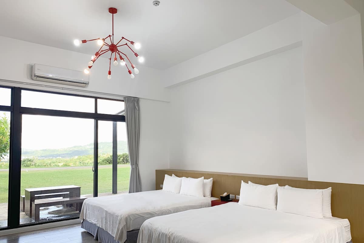 Una habitación de hotel con dos camas, un ventanal amplio con vistas a un paisaje verde, un equipo de aire acondicionado y una lámpara de araña roja singular.