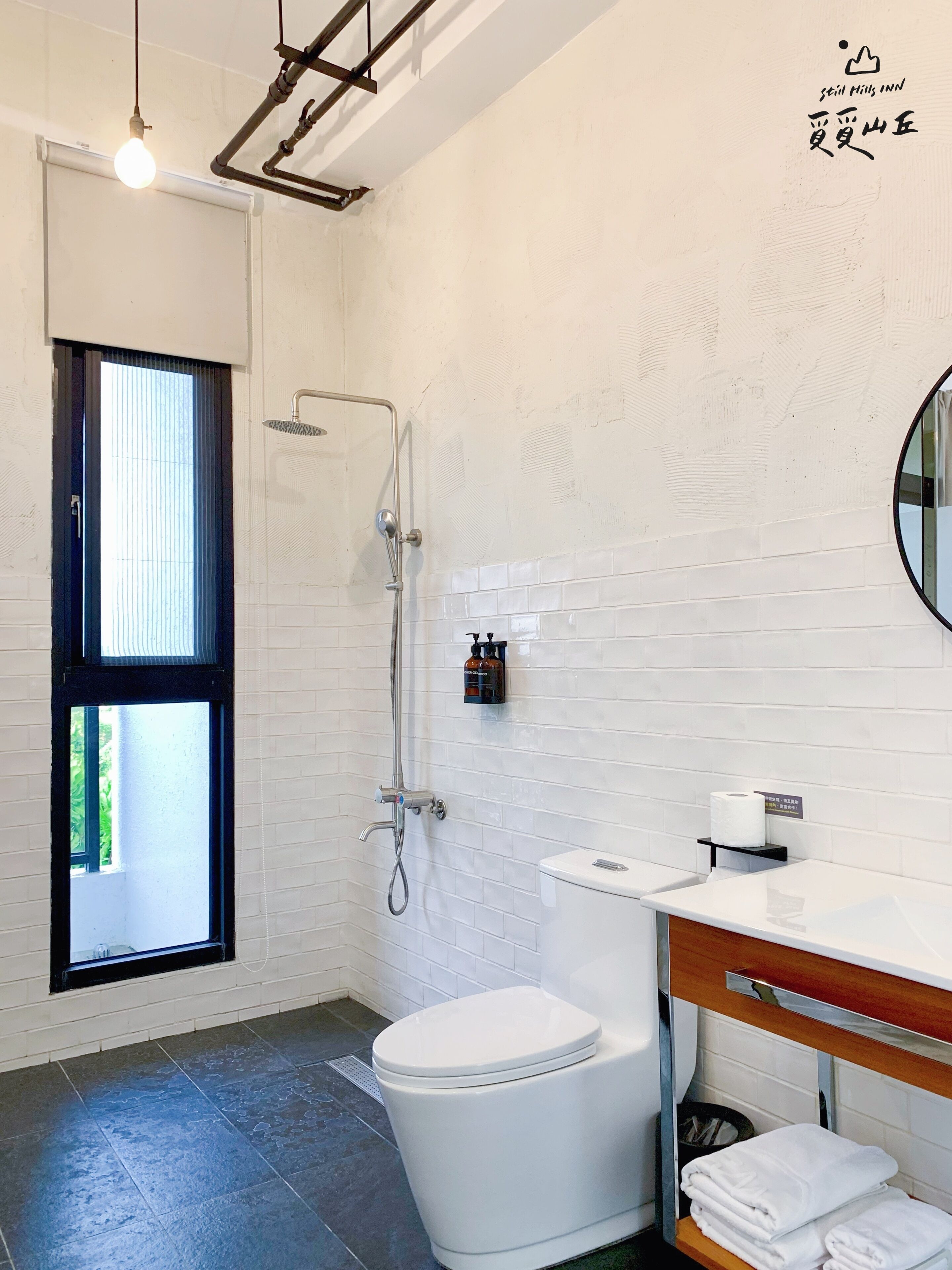 Un baño moderno con inodoro blanco, ducha y lavabo. Hay una ventana que proporciona luz natural y un perchero con toallas.
