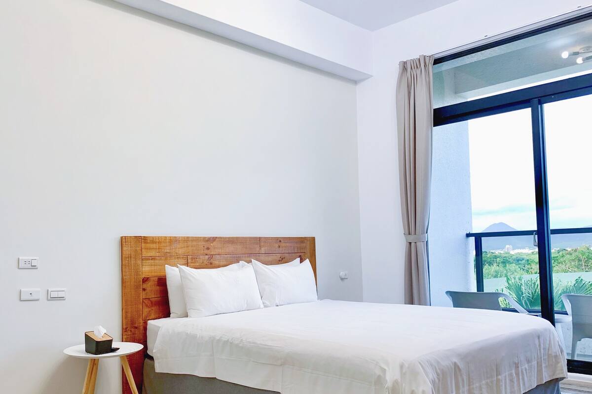 Un dormitorio moderno con cabecera de madera, una cama grande, una pequeña mesa redonda con un cofre y vistas a la vegetación a través de un amplio ventanal.