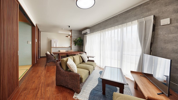 Ein modernes Wohnzimmer mit einer Couch, einem Couchtisch und einem Fernseher. Es gibt einen Essbereich mit Tisch und Stühlen. Der Raum hat Holzböden und ein großes Fenster mit Vorhängen.