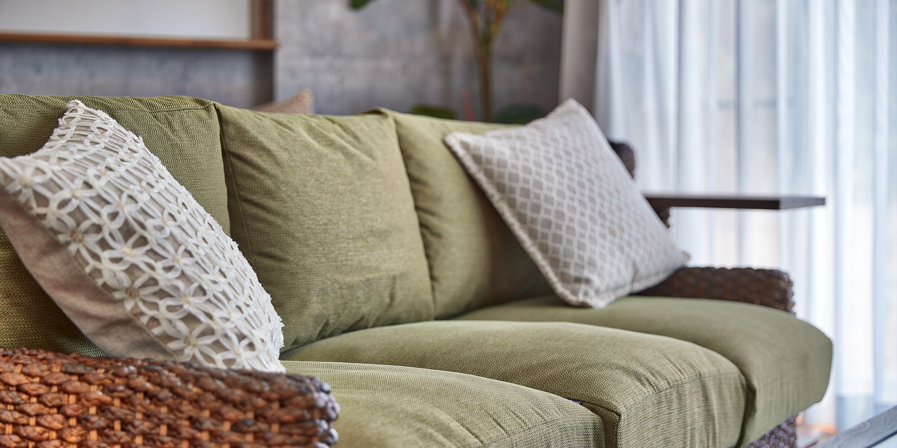 A green sofa with white and patterned cushions, positioned in a room with a window and a plant in the background.