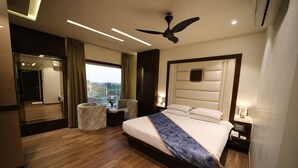 A modern hotel room with a large bed, a ceiling fan, a nightstand, a bedside table, and a view of the outdoors. - Hotel Prince Viraj (Jabalpur)