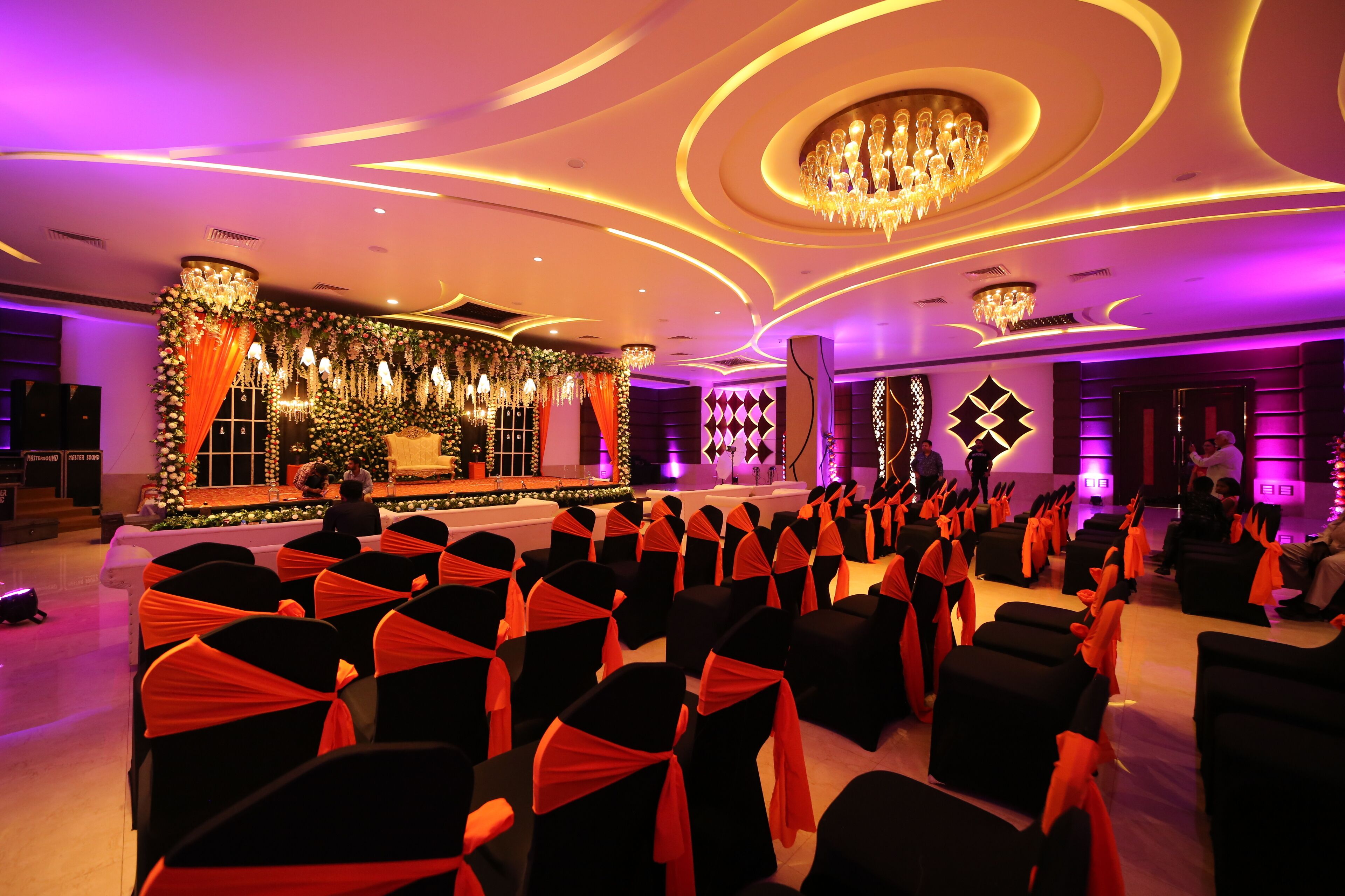 Un salón de banquetes con filas de sillas, un escenario con decoración floral y una araña de cristal.