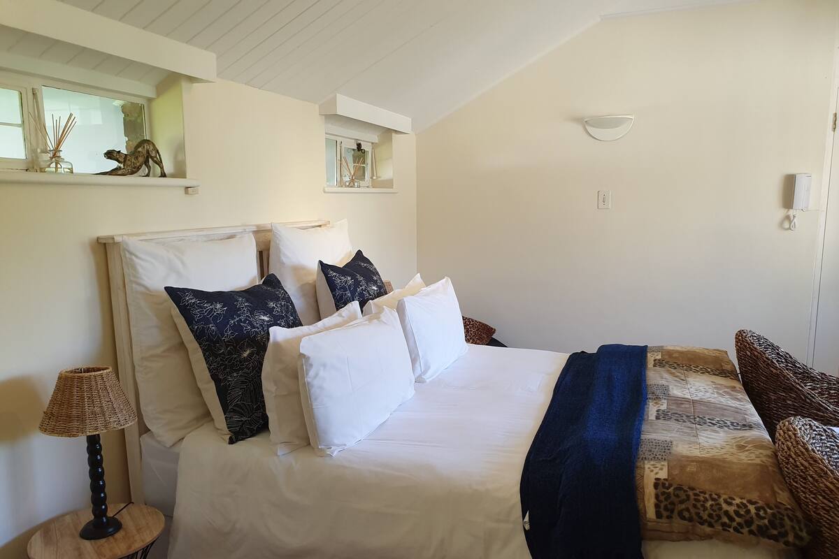 A neatly made bed with white and dark blue pillows, a blue throw blanket, and a woven chair.