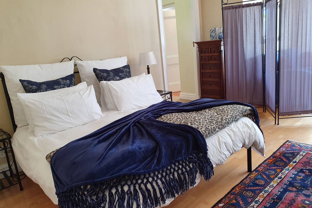 A neatly made bed with white and blue pillows, a blue blanket, and a patterned throw. A wooden nightstand with a lamp is beside the bed. A wooden dresser and a purple curtain are visible in the background.