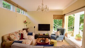 A living room with a sofa, armchair, coffee table, TV, and large windows with a view of greenery. - On Pinewood Guest House (Cape Town)