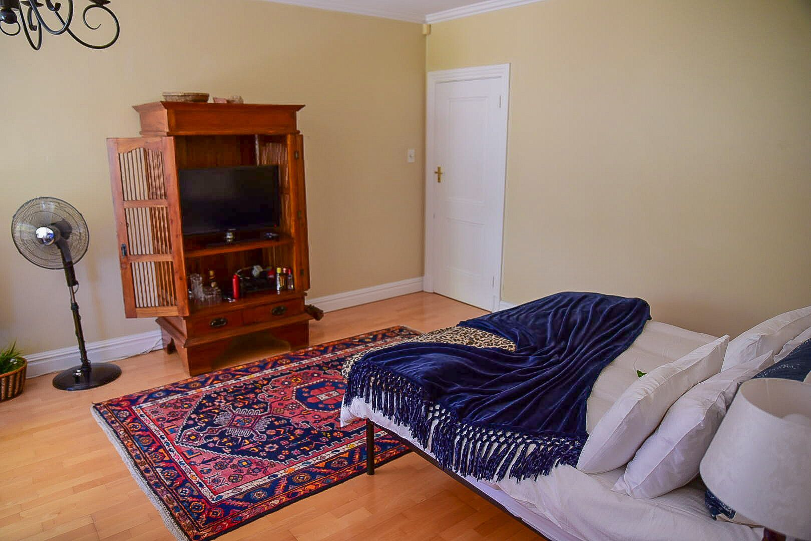 A room with a bed, a wooden entertainment unit, a standing fan, and a patterned rug.