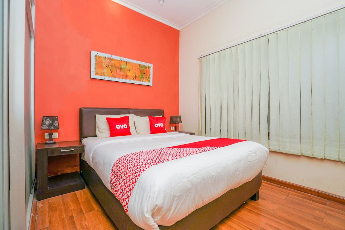 A hotel room with a large bed, red and white bedding, a nightstand with a lamp, a painting on the wall, and a window with blinds.